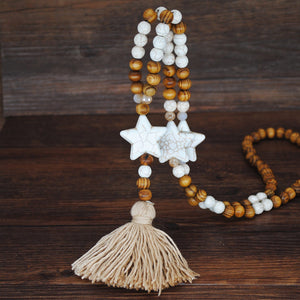 A long, beaded necklace with round, golden hued wooden beads and white "crackle glazed" beads, interspersed with three, large, white stars. The necklace culminates with light gold tassel pendant.