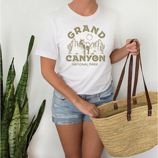 Woman with large straw bag wearing jean shorts and a short sleeved, crew neck t-shirt with a graphic that says "Grand Canyon National Park" around a canyon scene with cacti, a donkey, and a circle sun/moon.